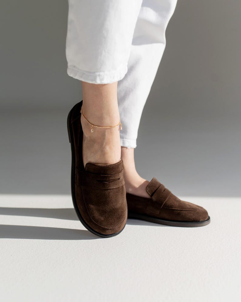 May include: A pair of dark brown suede loafers featuring a classic penny slot design across the vamp, a rounded toe, and a flat black sole. The shoes are worn with white trousers and a delicate gold chain anklet with small dangling charms.