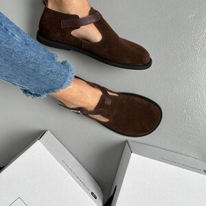 May include: Brown suede Mary Jane style shoes with a T-strap and a buckle closure. The shoes have a rounded toe and a flat sole. The shoes are displayed on a gray surface.