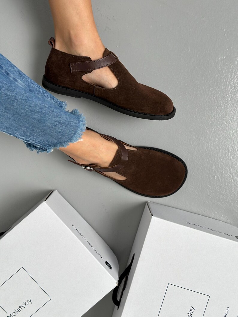 May include: Brown suede Mary Jane style shoes with a T-strap and a buckle closure. The shoes have a rounded toe and a flat sole. The shoes are displayed on a gray surface.