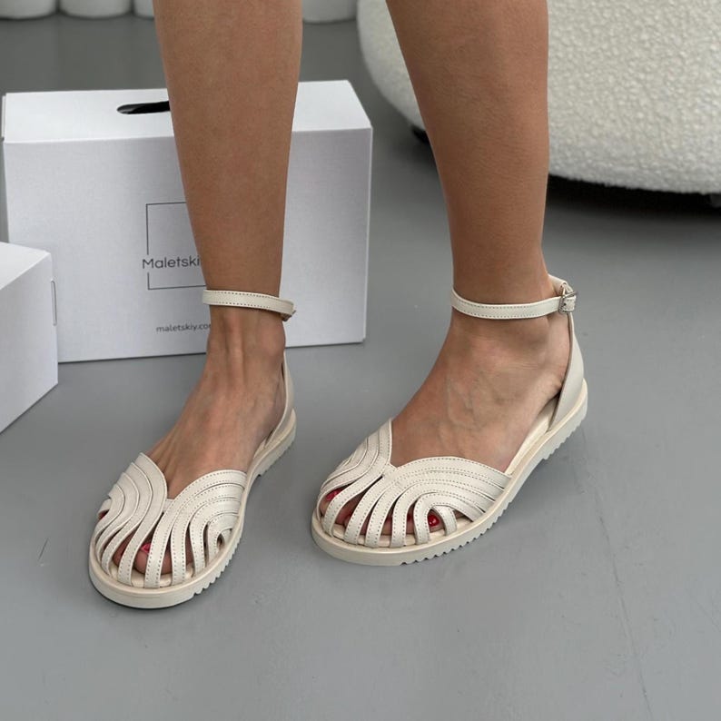 Puede incluir: Sandalias de cuero color crema con suela plana y correa en el tobillo. Las sandalias presentan un dise&ntilde;o &uacute;nico con cortes curvos en la parte superior. La imagen tambi&eacute;n muestra una caja blanca con el texto "Maletskiy" y una direcci&oacute;n web.