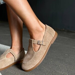 May include: Tan suede Mary Jane style flats with a buckle closure. The shoes have a rounded toe and a brown sole. The shoes are a neutral color and could be worn with many outfits.