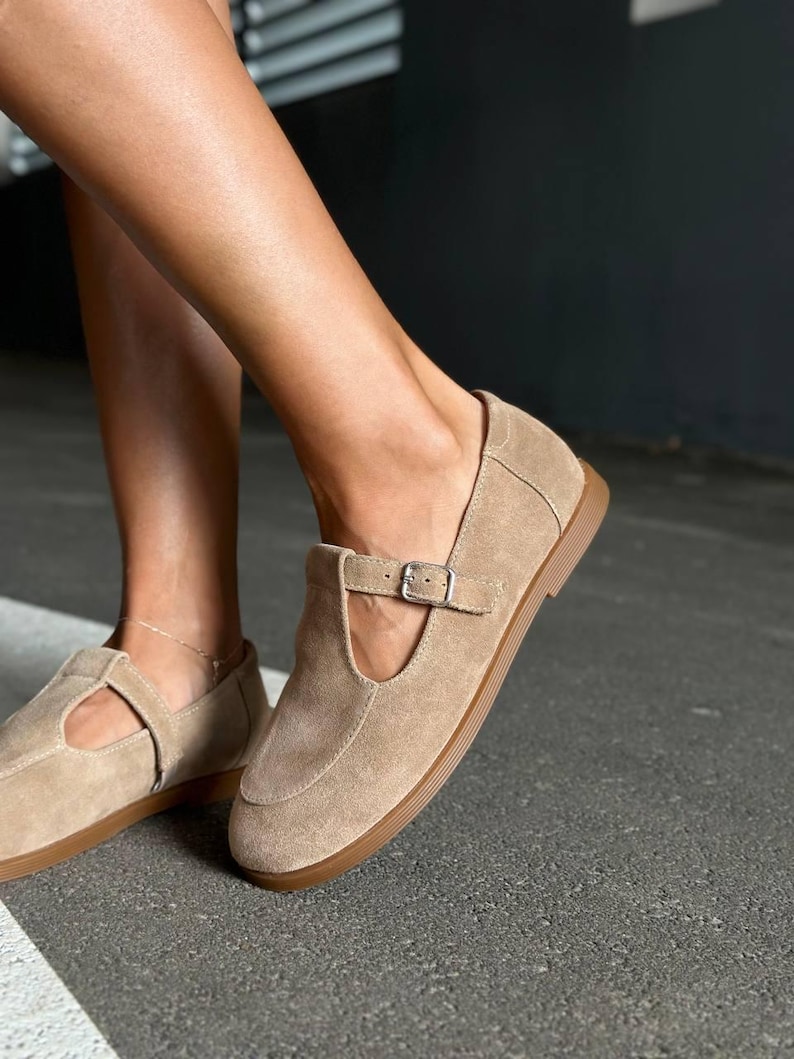 May include: Tan suede Mary Jane style flats with a buckle closure. The shoes have a rounded toe and a brown sole. The shoes are a neutral color and could be worn with many outfits.