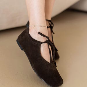 May include: Close-up of brown suede flats with a square toe and tie-up laces. The shoes have a low heel and a cutout detail on the side. A silver ankle bracelet is visible.
