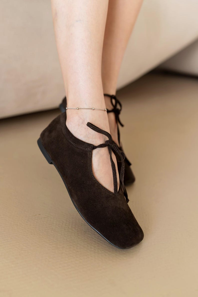 May include: Close-up of brown suede flats with a square toe and tie-up laces. The shoes have a low heel and a cutout detail on the side. A silver ankle bracelet is visible.