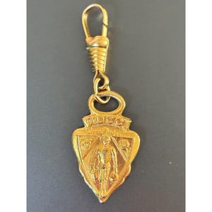 May include: A gold-toned metal charm featuring a shield-shaped pendant with a knight figure embossed in the center. The word GUCCI is engraved at the top of the shield. The pendant is attached to a ribbed lobster claw clasp by a circular jump ring.