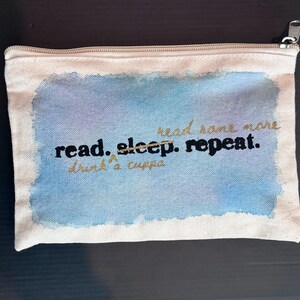 May include: A rectangular beige canvas zippered pouch featuring a watercolor-style blue and purple rectangular background. The text reads read, sleep, repeat in black, with read some more and drink a cuppa written in gold cursive script.