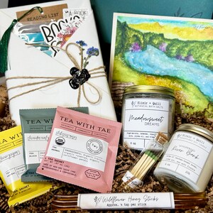 May include: A gift box filled with self-care items. Includes a wrapped book, watercolor painting, tea sachets, bath salts, a candle, matches, and honey sticks. The box is lined with brown crinkle paper.