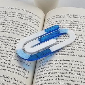 Puede incluir: Un libro abierto con un marcapáginas azul y blanco. Una pequeña planta suculenta en una maceta blanca está a la izquierda del libro. El marcapáginas tiene un diseño en forma de U con detalles azules y blancos.