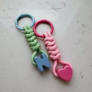May include: Two keychains featuring braided nylon cord. One has a light blue circular clasp, light green cord, and a blue letter N charm. The other has a pink circular clasp, pink cord, and a pink heart-shaped charm attached to the bottom.
