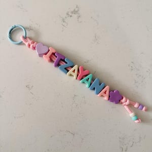 May include: A keychain featuring a light blue circular clasp attached to a pink cord. The cord is threaded with multicolored letter beads spelling ITZAYANA, interspersed with a purple heart bead at the start and a purple flower bead at the end.