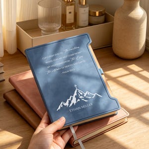May include: A blue leather-bound journal with a tabbed edge, featuring a white mountain graphic, the name "Ethan Miller," and the text "I lift up my eyes to the mountains. Where does my help come from? My help comes from the Lord."