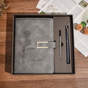 May include: A gray journal with a floral design and the name "Amelia" is displayed in a gift box. The journal has a gold-colored clasp and comes with a pen and a pen refill. The journal and pen set is on a wooden surface.