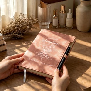 May include: A dusty rose-coloured notebook with a white floral line drawing and the text "THE NEXT chapter SAMUEL" printed on the cover. The notebook features a black pen held in a side loop and a silver ribbon bookmark, resting on a wooden surface.