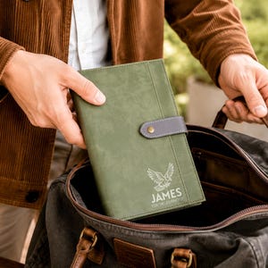 May include: An olive green notebook with a grey snap closure and a white eagle graphic above the text JAMES FOR THE JOURNEY. The notebook is being placed into a dark grey canvas bag with brown leather accents and handles.