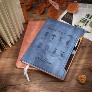 May include: A blue journal with the text "GOD SAYS YOU ARE" and floral illustrations. A pen is inserted in the side and a white ribbon bookmark is visible. A peach-coloured journal is behind the blue one.