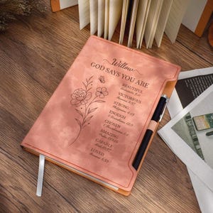 Op de afbeelding: Een perzikroze notitieboek met de tekst "Willow GOD SAYS YOU ARE" en inspirerende woorden. Een bloemenontwerp en een pen zijn zichtbaar. Het notitieboek heeft een lint-bladwijzer.