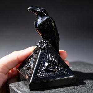 Op de afbeelding: Een sculptuur van gepolijste zwarte steen met een raaf bovenop een piramide. De piramidevormige basis toont een uitgesneden oogsymbool op de zichtbare zijde, met gedetailleerde stralen die uit het oog komen. Het object heeft een gladde afwerking.