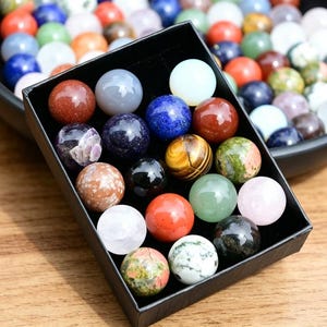 May include: A selection of polished gemstone spheres in a variety of colours, including red, blue, green, and white. Some spheres are in a black box, while others are in a black bowl. The spheres are displayed on a wooden surface.