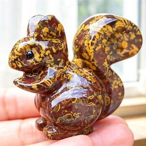 Puede incluir: Una talla de piedra pulida con forma de ardilla que presenta un color base marrón rojizo intenso con intrincados patrones arremolinados en amarillo y dorado, incluyendo una cola grande y curva y una cabeza pequeña y redondeada con ojos negros.