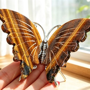 Könnte beinhalten: Ein schmetterlingsförmiges Ornament aus poliertem Tigerauge-Edelstein mit braunen und goldenen Streifenmustern. Die Flügel sind mit feinen Linien graviert und der Mittelkörper besteht aus silberfarbenem Metalldraht.