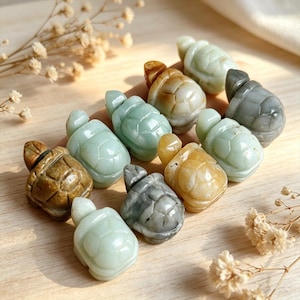 May include: A collection of carved stone turtle figurines in various colors, including green, brown, and gray. The turtles have detailed shell patterns and are arranged on a light-colored surface. Some have a polished, glossy finish.