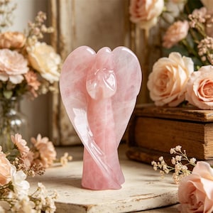 Pode incluir: Uma estatueta de anjo de quartzo rosa esculpida com asas abertas. O anjo é de uma cor rosa suave e está sobre uma superfície branca. O fundo inclui flores e um livro de madeira vintage.