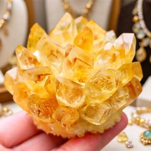 Op de afbeelding: Een cluster van gouden citrienkristallen, met een stralende, honingkleurige tint. De edelsteenformatie wordt in een hand gehouden, met diverse gouden sieraden op de achtergrond, waaronder kettingen, ringen en armbanden.