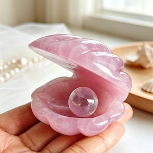May include: A carved light pink rose quartz seashell sculpture featuring a hinged design that opens to reveal a smooth, spherical, translucent light purple crystal ball resting inside the center of the polished stone shell structure.