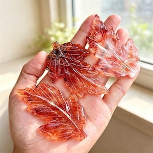 Peut inclure: Trois sculptures translucides en forme de feuille, taillées dans une pierre rouge orangé avec des inclusions et des nervures internes visibles. Chaque pièce présente un bord dentelé détaillé et une pointe, imitant une feuille d'érable.