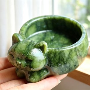 May include: A small, polished green stone bowl carved into the shape of a resting cat. The bowl features a smooth, rounded interior and a detailed cat head with closed eyes and pointed ears extending from the front edge of the circular vessel.