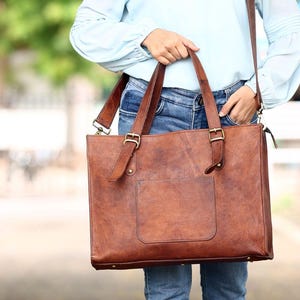 May include: A rectangular brown leather tote bag with two top handles, adjustable buckle straps, and a front pocket. The bag features a smooth, distressed finish and metallic hardware, designed for carrying items with a structured, spacious interior.