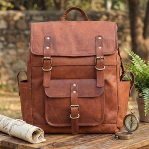 Pode incluir: Uma mochila de couro castanho com uma aba superior grande com duas correias de fivela, um bolso frontal com uma correia de fivela, dois bolsos laterais e uma pega superior. O saco apresenta costuras visíveis e ferragens em tom de latão.