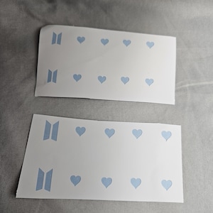 Op de afbeelding: Twee witte stickerbladen met lichtblauwe hart- en logo-uitsparingen. Elk blad heeft een rij van vier harten en twee logo-ontwerpen. De stickers liggen op een grijze ondergrond.