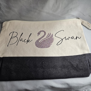 May include: A canvas zipper pouch with a black base and a cream-colored upper section. The words "Black Swan" are printed in a script font, with a glittery purple swan graphic. The pouch has a zipper closure.