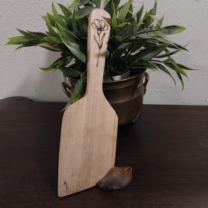 May include: A light-colored wooden paddle with a long handle featuring a dark, thin-lined botanical illustration of a single flower etched into the wood. The paddle has a flat, wide base and a smooth, natural grain finish throughout the surface.
