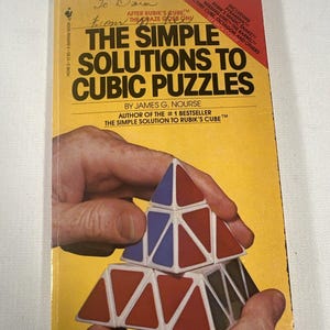 May include: A paperback book titled The Simple Solutions to Cubic Puzzles by James G. Nourse. The yellow cover features a graphic of a triangular puzzle with red, blue, and white segments. Handwritten ink text appears at the top of the cover.