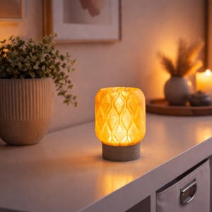 May include: A lit, decorative lamp with a geometric pattern. The lamp has a warm, yellow glow and sits on a gray base. The lamp is on a white surface, with a potted plant and other decor in the background.