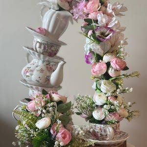 May include: A decorative arrangement featuring a vertical stack of white ceramic teacups and a teapot, adorned with an arch of artificial pink, white, and purple flowers. A single floral-patterned teacup sits at the base on a wooden slice.