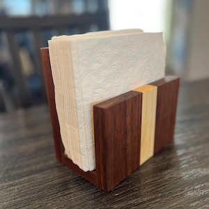 Puede incluir: Un servilletero de madera en color marrón oscuro, con una franja de madera más clara. Contiene una pila de servilletas blancas estampadas. El diseño es sencillo y moderno, adecuado para una mesa de comedor o cocina. Mide unos 15 cm x 15 cm.