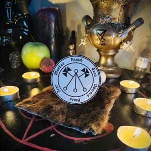 May include: A white circular sticker with black text and symbols, reading "Marbas, Renich, Tasa, Liberaca, Biasa, Jcar". The sticker is on a brown fur-like surface, surrounded by lit candles and a golden urn.