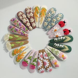 May include: A collection of embroidered hair clips in various colors and floral designs. The clips feature detailed stitching of flowers, leaves, and ladybugs on a variety of pastel and neutral backgrounds. The clips are arranged in a circular pattern.
