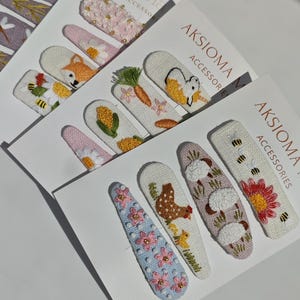 May include: A collection of hair clips mounted on white cards labeled AKSIOM ACCESSORIES. The clips feature embroidered designs including flowers, a fox, a carrot, a unicorn, a chicken with chicks, sheep, and bees in various colors.