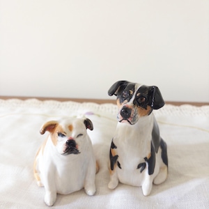 May include: Two ceramic dog figurines. One is a white and tan bulldog with a slightly closed eye. The other is a black, white, and tan terrier mix. Both are sitting on a white cloth.