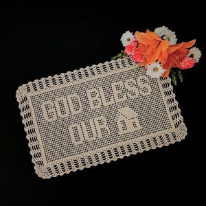 May include: Cream-colored, crocheted rectangular doily with the words "GOD BLESS OUR" and a house design. The doily has a scalloped edge and is decorated with orange and white flowers.