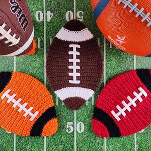 May include: Four crocheted footballs in brown, orange, red, and white. The footballs are on a green and white football pitch.