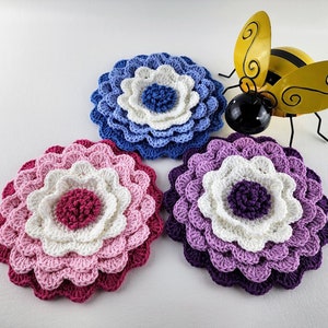 Puede incluir: Tres posavasos de ganchillo con forma de flor en tonos de rosa, morado y azul. Los posavasos están superpuestos con diferentes tonos de cada color, creando un efecto multidimensional. Una figurita de abeja amarilla y negra se coloca en el centro de la imagen.