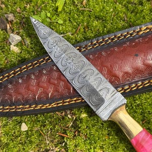 Pu&ograve; includere: Un coltello in acciaio Damasco con lama a fantasia, rinforzo in ottone e manico rosa. Il coltello poggia su un fodero in pelle marrone con cuciture decorative. La lama misura circa 20 cm.