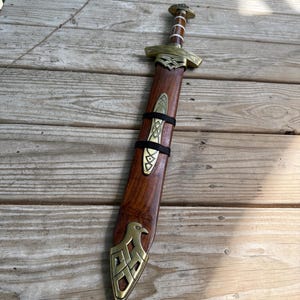 May include: A sword with a wooden scabbard featuring brass fittings. The scabbard has a brass eagle motif at the tip and a central brass plate secured by black straps. The sword hilt is made of wood with brass accents and a rounded pommel.