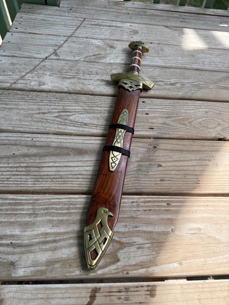 May include: A sword with a wooden scabbard featuring brass fittings. The scabbard has a brass eagle motif at the tip and a central brass plate secured by black straps. The sword hilt is made of wood with brass accents and a rounded pommel.