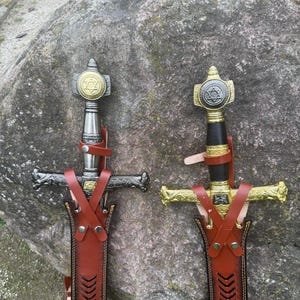 May include: Two decorative swords with ornate hilts and crossguards are displayed. The hilts feature a gold and silver design with a star emblem. The swords are sheathed in brown leather scabbards with decorative stitching.